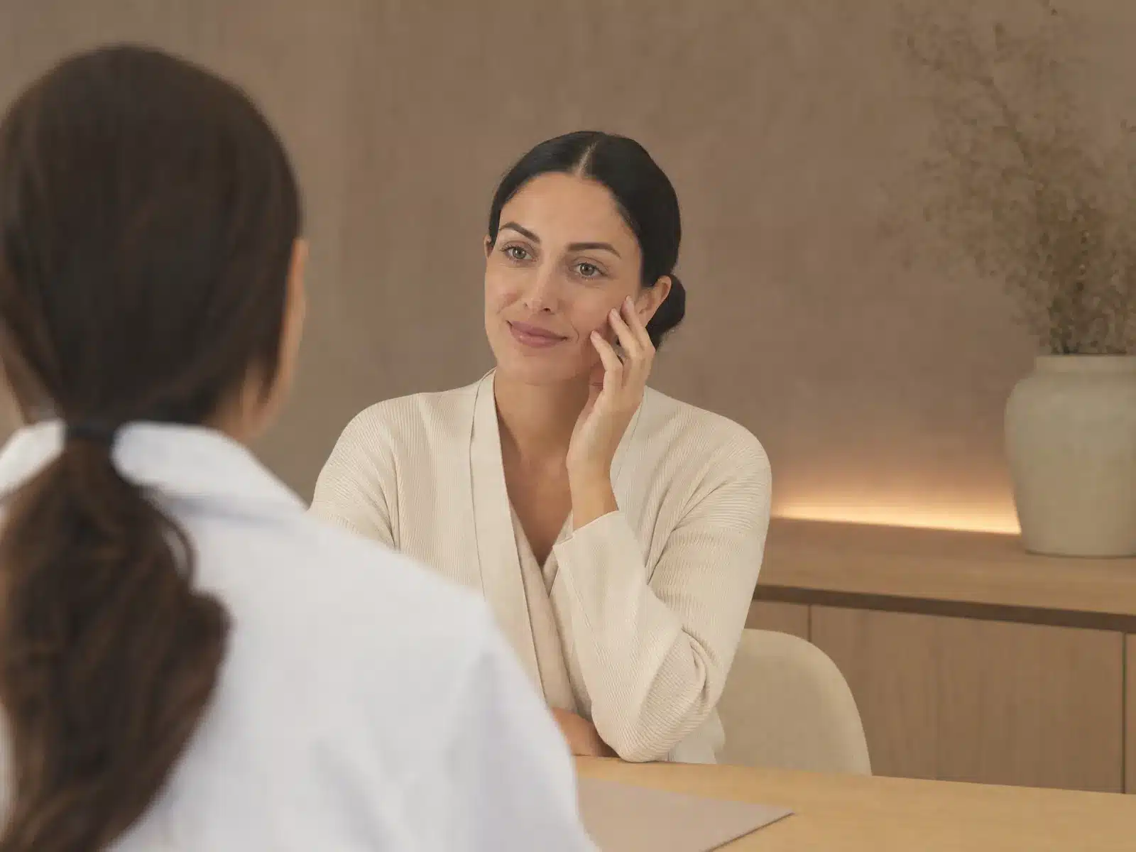 Consultation avant une injection d'acide hyaluronique : femme souriante écoutant un médecin en blouse blanche, la main posée sur la pommette