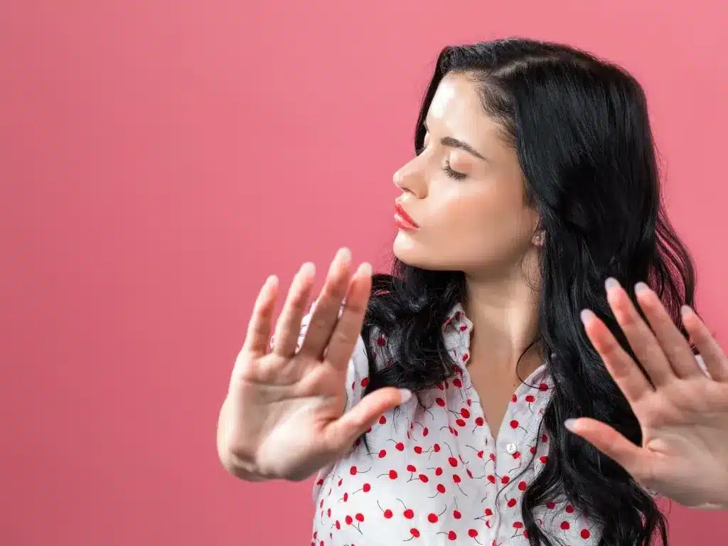 Contre-indications de l'injection d'acide hyaluronique : femme brune en chemisier à pois tendant les mains pour dire stop, sur fond rose