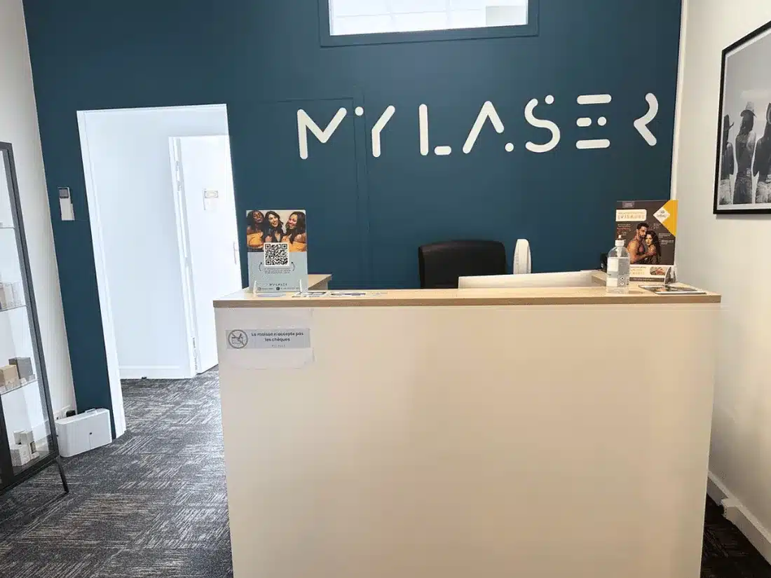 Accueil de My Laser Bordeaux : banque blanche au plateau bois devant un mur bleu foncé portant le logo MYLASER en lettres blanches, vitrine vitrée à gauche