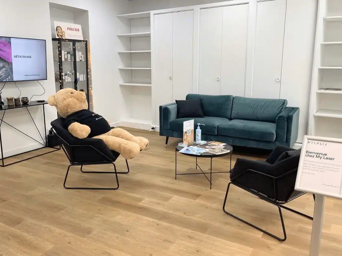 Salle d'attente de My Laser Nantes : canapé en velours bleu canard, fauteuils noirs, grand ours en peluche en tee-shirt My Laser et écran qui présente le détatouage