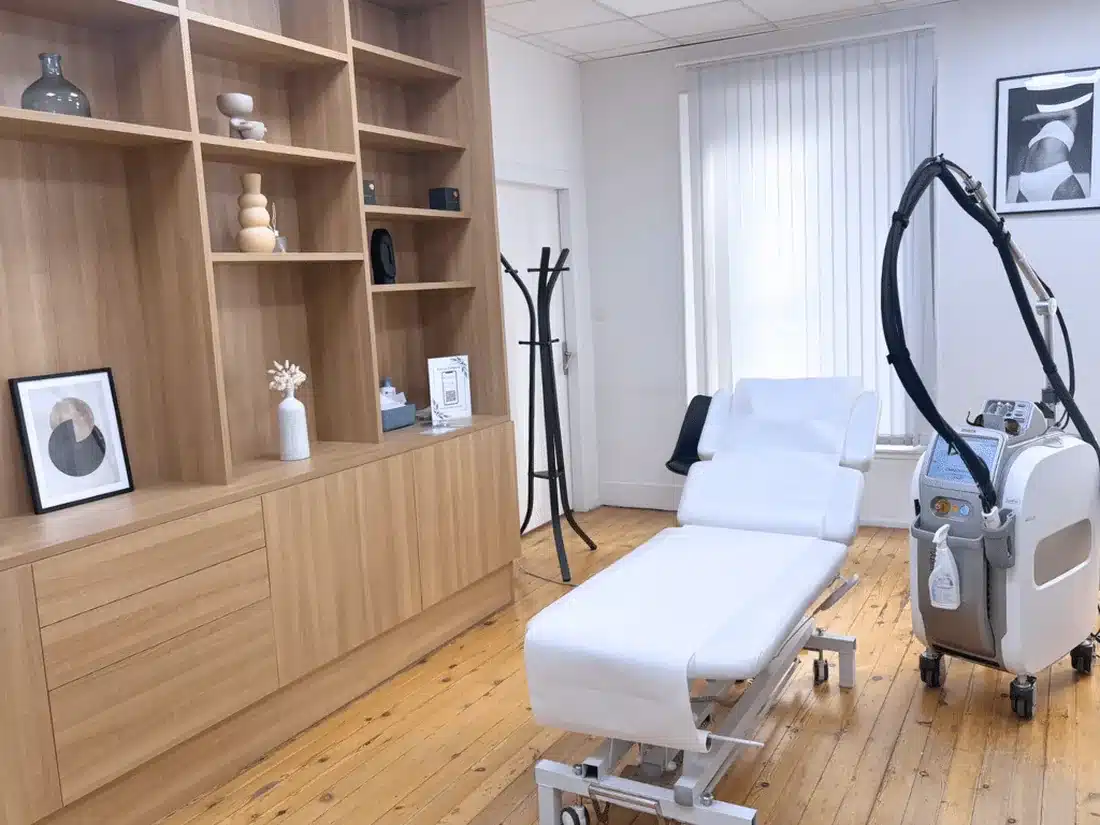 Salle de soin de My Laser Toulouse : table de soin blanche, laser d'épilation, étagères en bois clair et store vertical