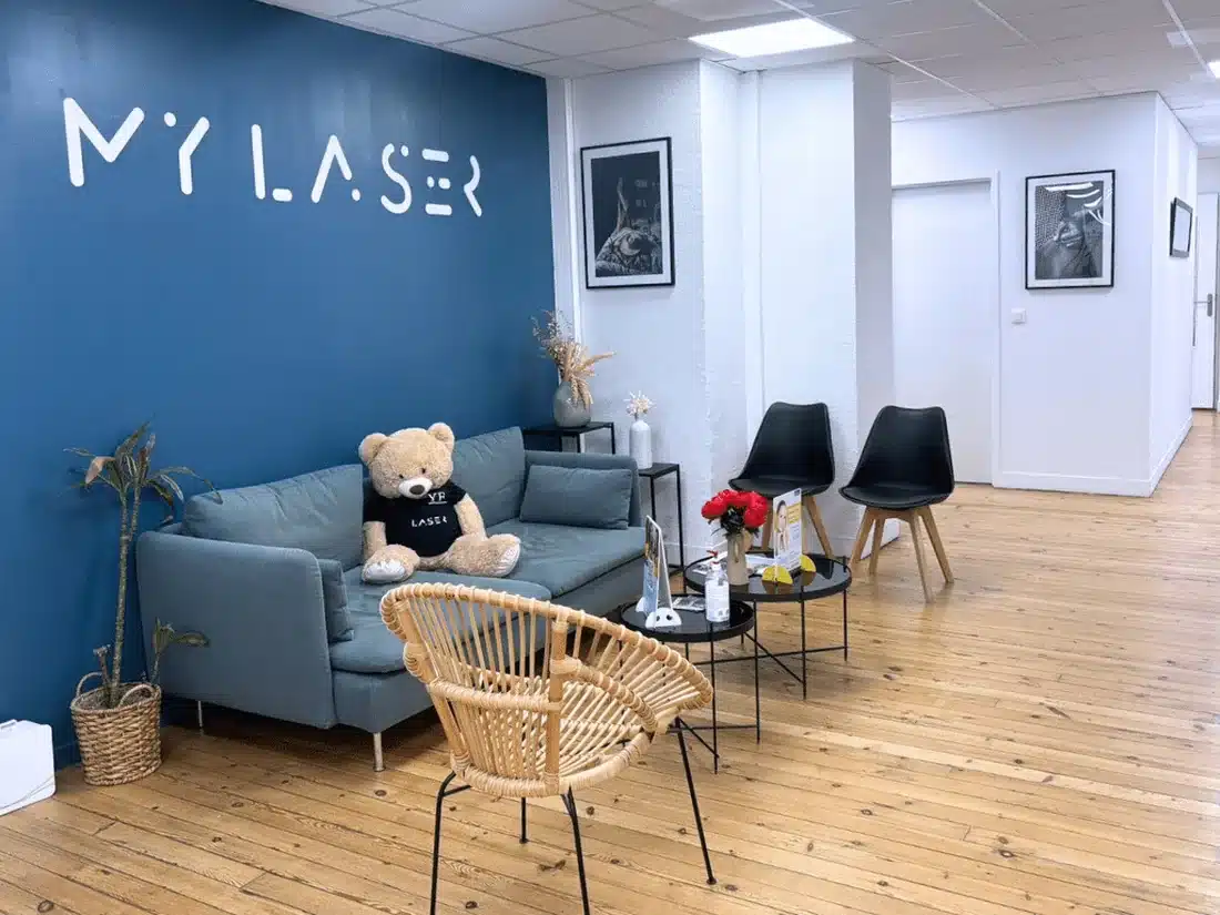 Accueil de My Laser Toulouse : mur bleu avec le logo My Laser, canapé gris-bleu et ours en peluche, fauteuil en rotin, deux chaises noires et parquet clair