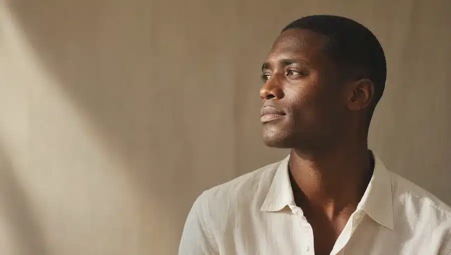 Portrait d'un homme à la peau brun foncé, mâchoire et cou rasés de près et lisses, chemise en lin ivoire au col ouvert, regard de côté