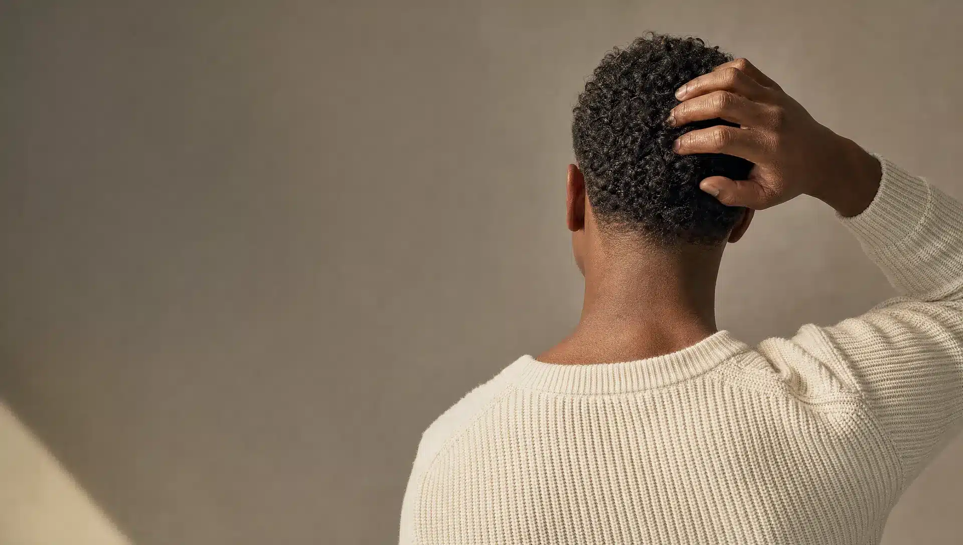Homme à la peau brun foncé vu de dos, la main dans ses cheveux courts, bouclés et denses, pull en maille ivoire