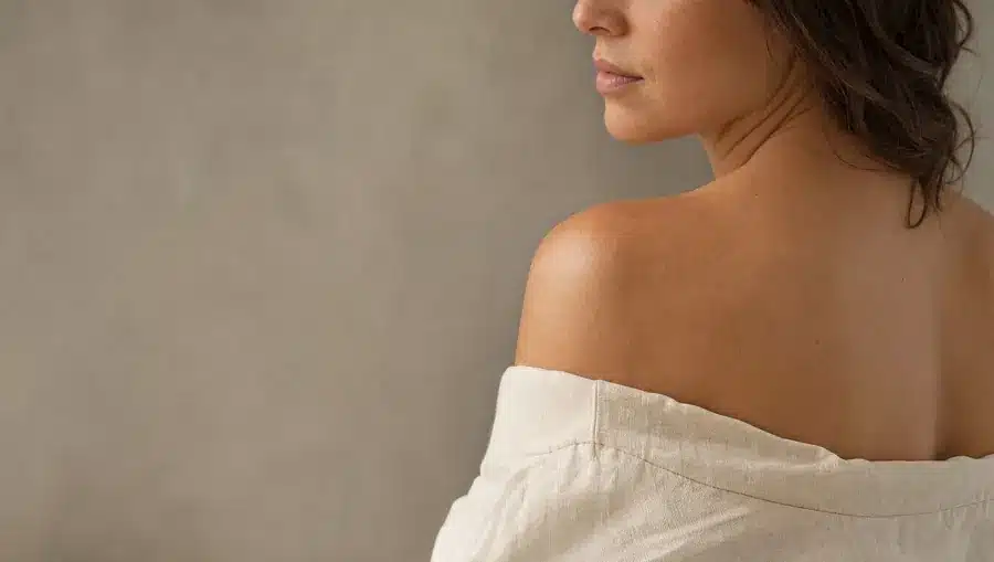 Épaule et haut du dos d'une femme à la peau légèrement hâlée avec quelques légères taches solaires, haut en lin ivoire glissé sur l'épaule