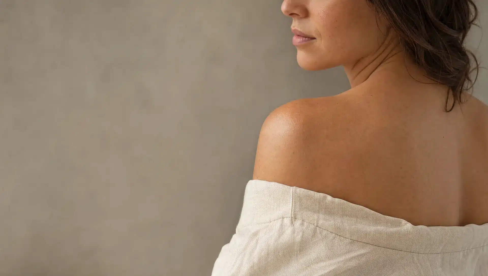 Épaule nue et haut du dos d'une femme de profil, peau légèrement hâlée avec quelques légères taches solaires, haut en lin ivoire descendu sous l'épaule