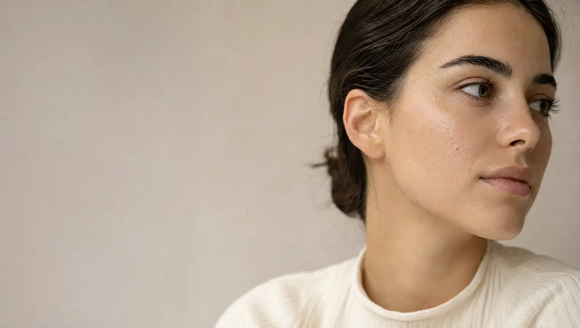 Jeune femme à la peau olive claire vue de trois quarts, quelques petites imperfections sur la joue, cheveux bruns attachés, pull ivoire côtelé
