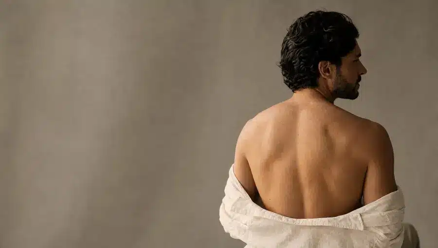 Homme à la peau olive claire vu de dos, haut du dos net et lisse, chemise en lin ivoire baissée à mi-dos