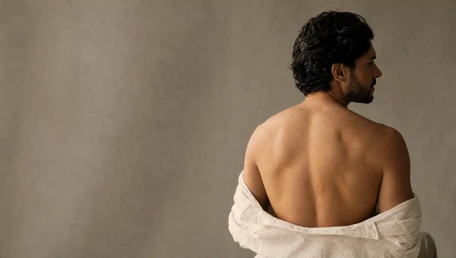 Homme à la peau olive claire vu de dos, haut du dos net et lisse, chemise en lin ivoire baissée à mi-dos