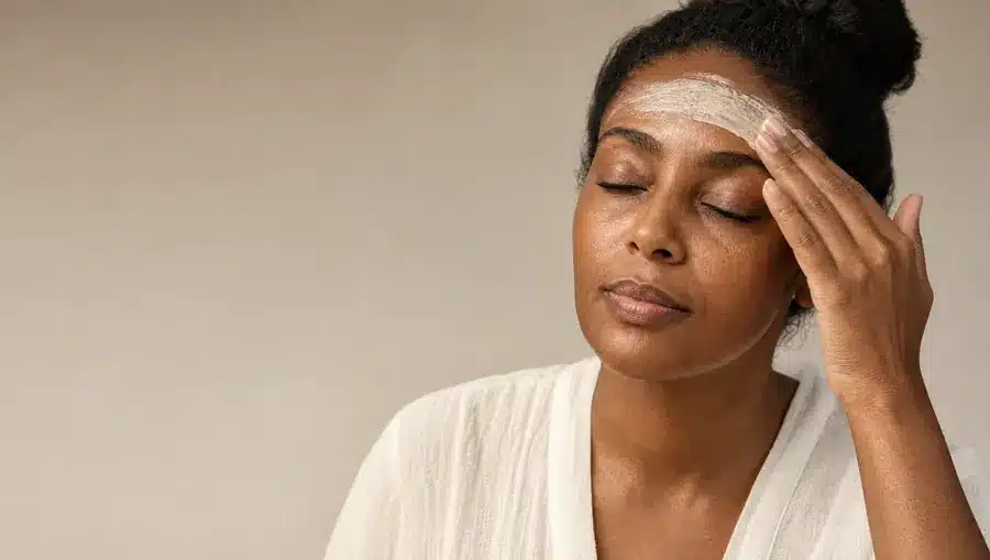 Femme à la peau brun foncé étalant du bout des doigts un masque crème ivoire sur son front, yeux fermés