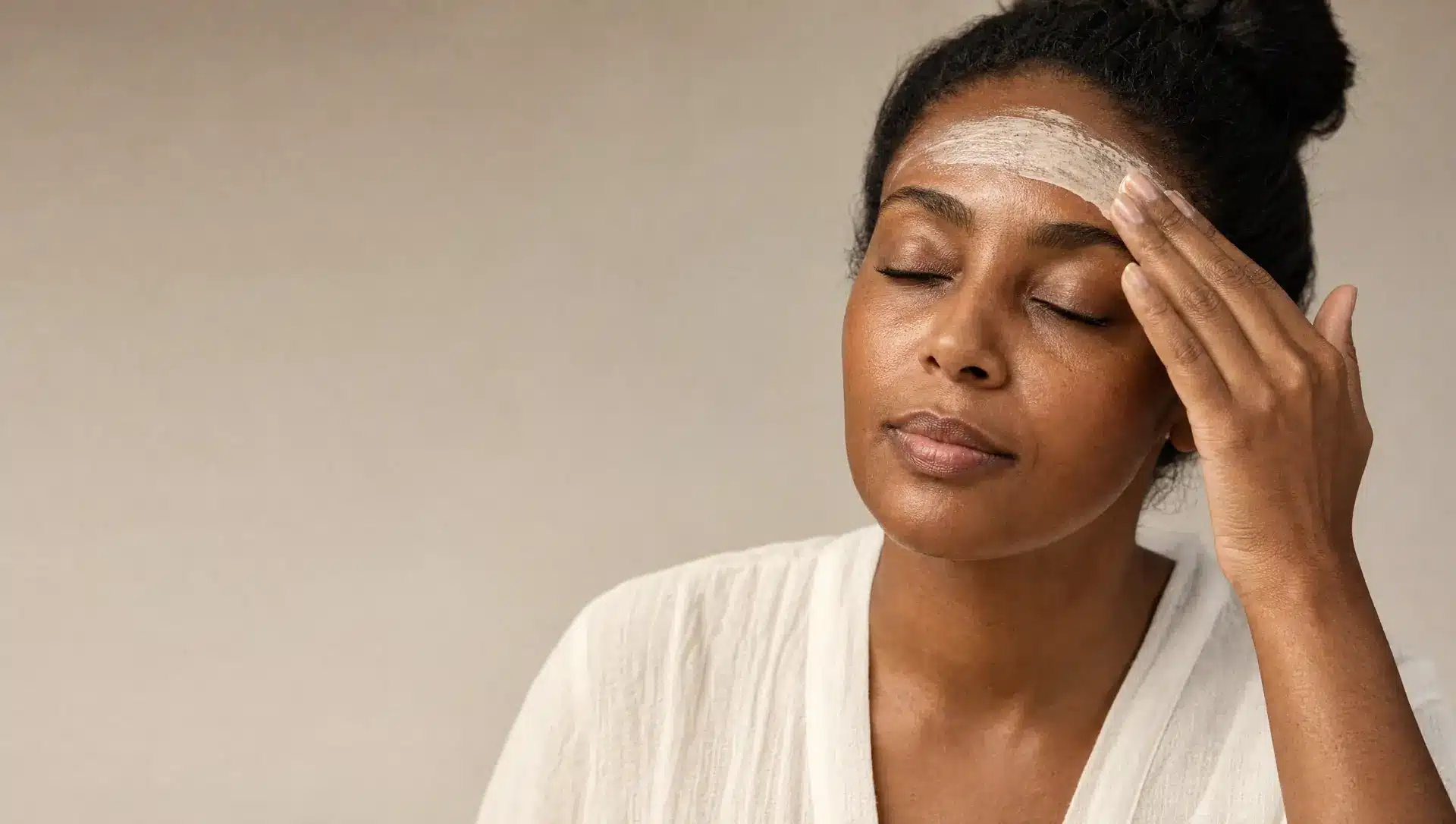 Femme à la peau noire, les yeux fermés, qui étale du bout des doigts un masque crème ivoire sur son front